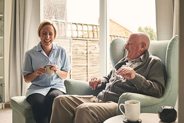 Indoor shot of smiling senior man and female carer enjoying coffee in living room