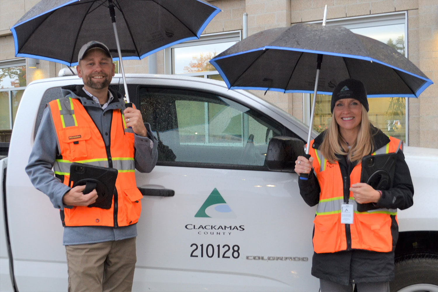 2 people with umbrellas in orange vest in front of county vehicle