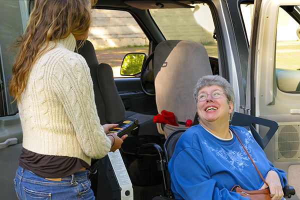 A disable woman in a wheelchair on a lift about to exit a handicapped accessible van, being helped by a young care giver.