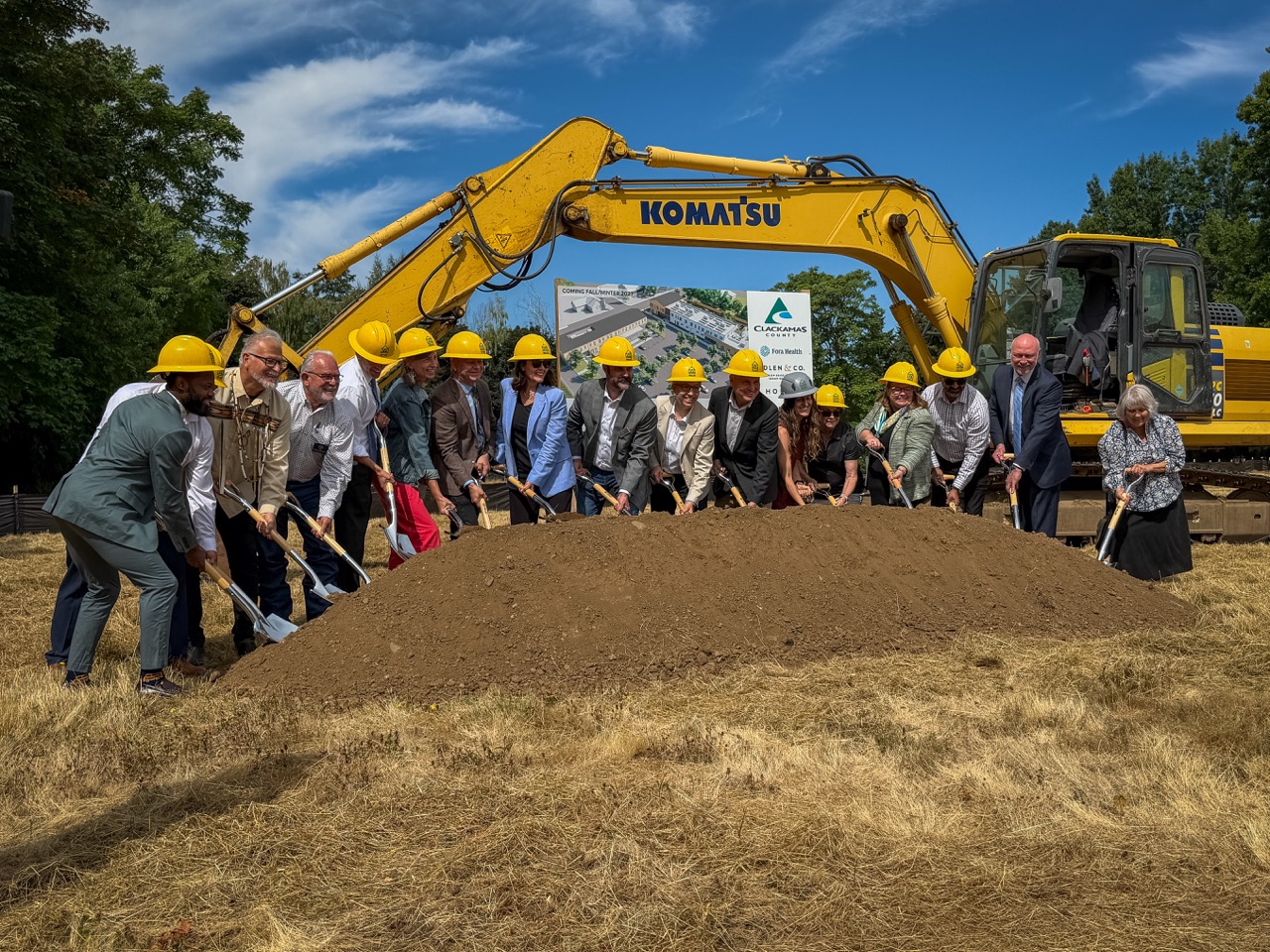 Elected officials shovel a dirt pile with a piece of construction equipment behind them, at the Clackamas County Recovery Campus groundbreaking on July 29, 2026. They are out in a field and all looking up toward a photographer. 