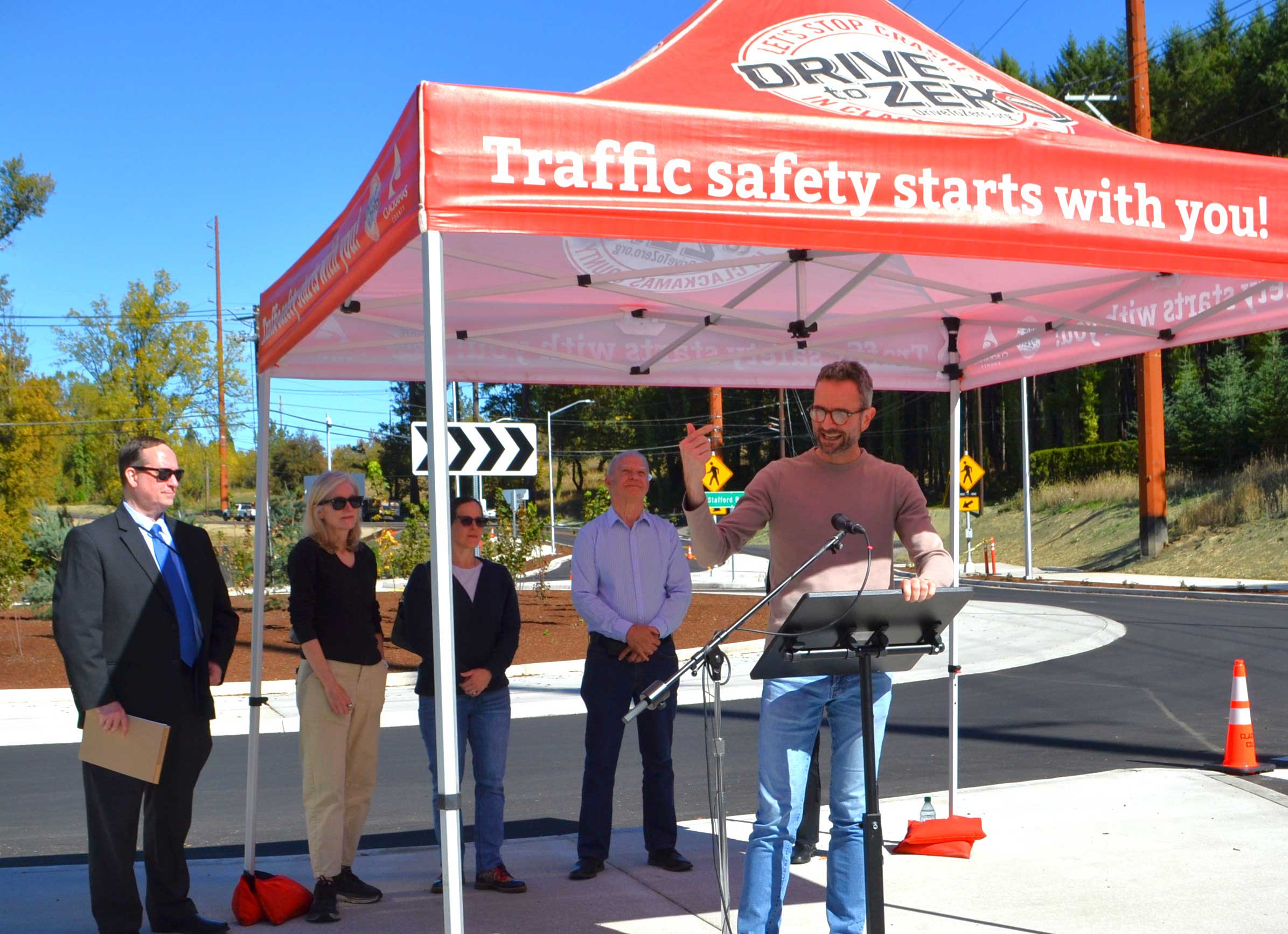 Lake Oswego Mayor Joe Buck speaks at the opening of the Stafford Roundabout on Sept. 30, 2026. Behind him are Clackamas County Administrator Gary Schmidt, Lake Oswego Councilors Trudy Corrigan and Rachel Verdick, and Clackamas County Commissioner Paul Savas. Above them is a Clackamas County Drive to Zero tent, with the words. Traffic safety starts with you!
