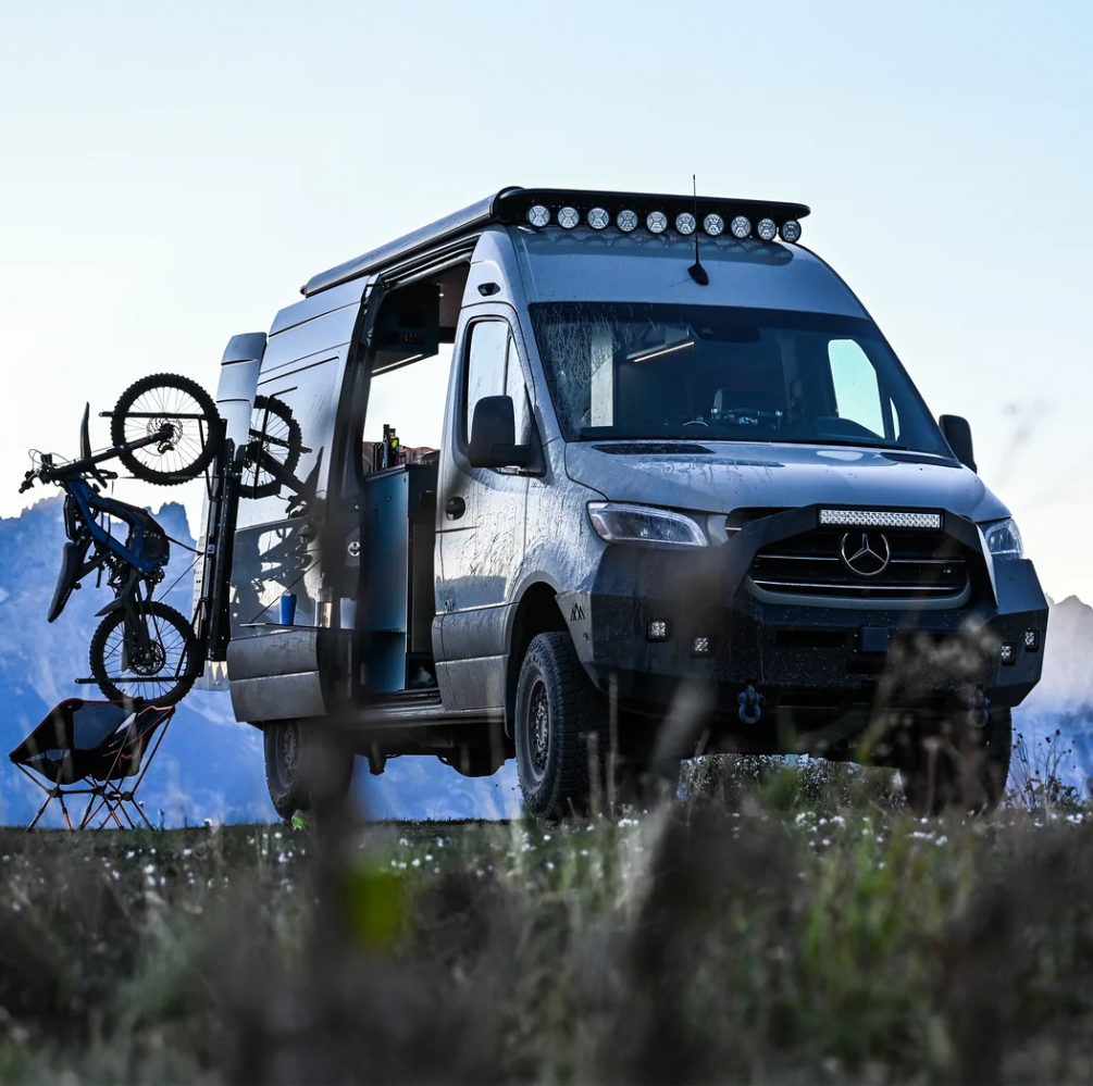 Large 4x4 RV with teh side door slid open and an ebike mounted to the back parked near mountains and wild flowers