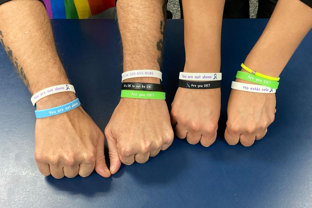 Two people hold out their wrists showing bracelets with encouraging messages that read, You are not alone, Are you okay?, and No estas solo.