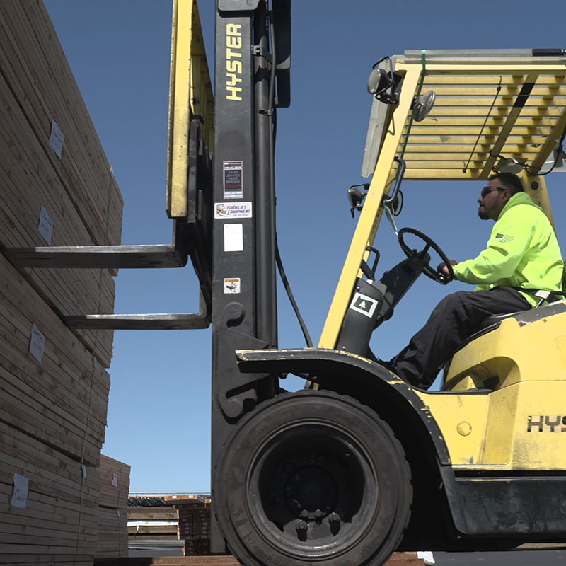 A worker operates a yellow forklift to move a tall stack of wooden planks in an outdoor industrial setting.