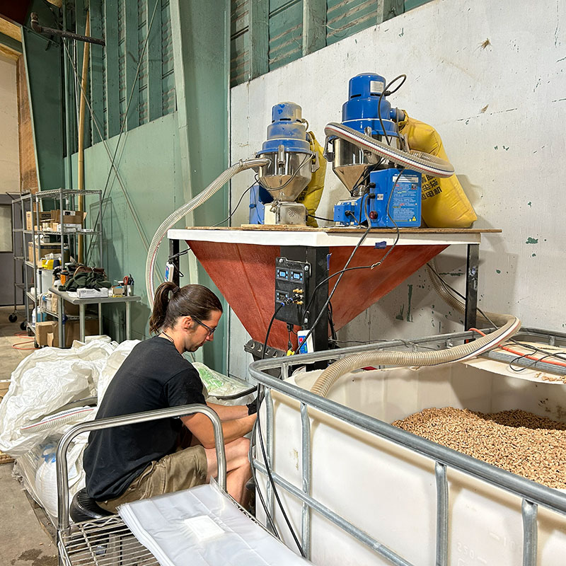 Man crouches in an industrial setting surrounded by plastic industrial tubes and there is an open vat of organic product next to him