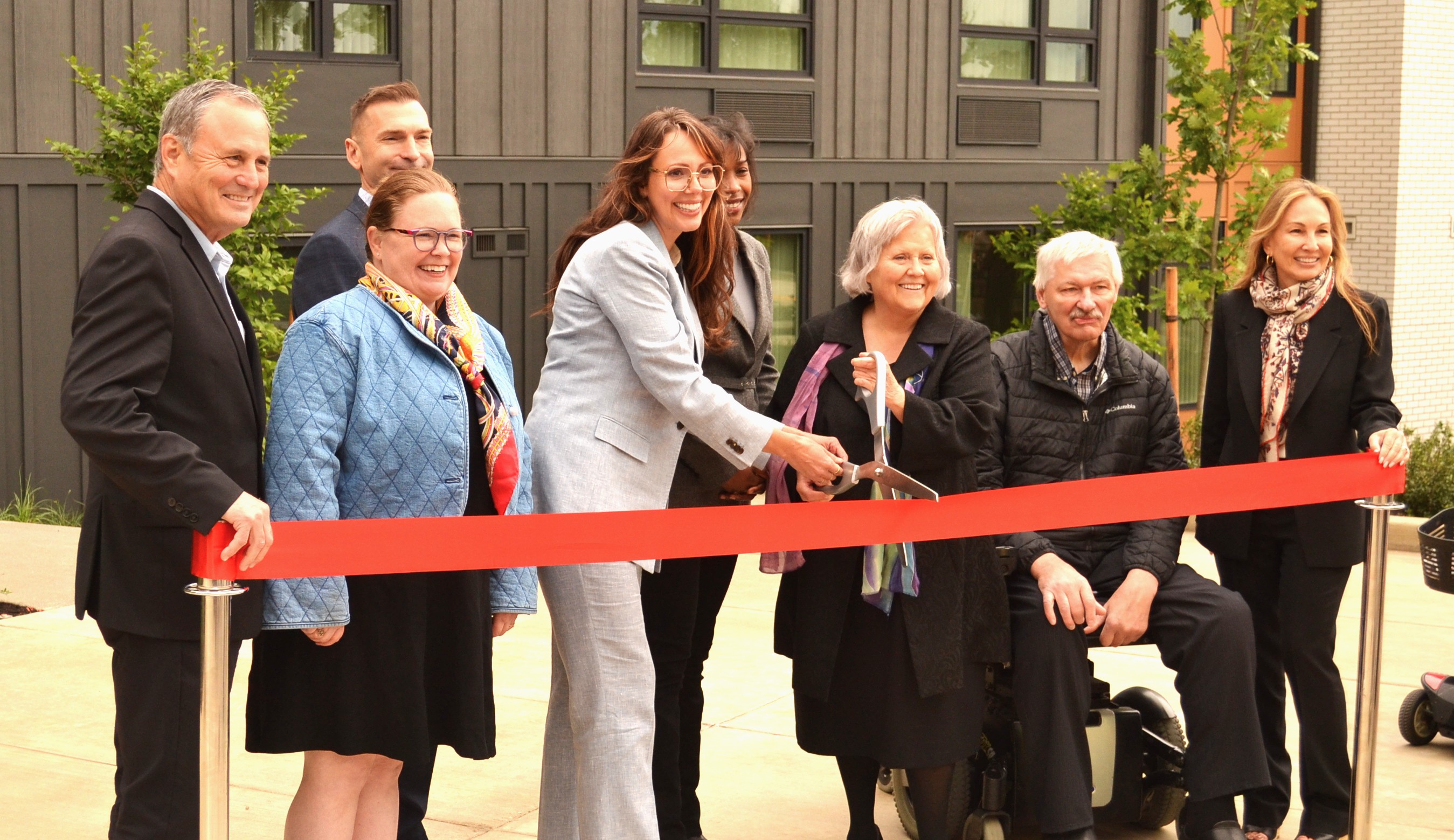 8 smiling people standing behind a red ribbon preparing to be cut with large scissors.