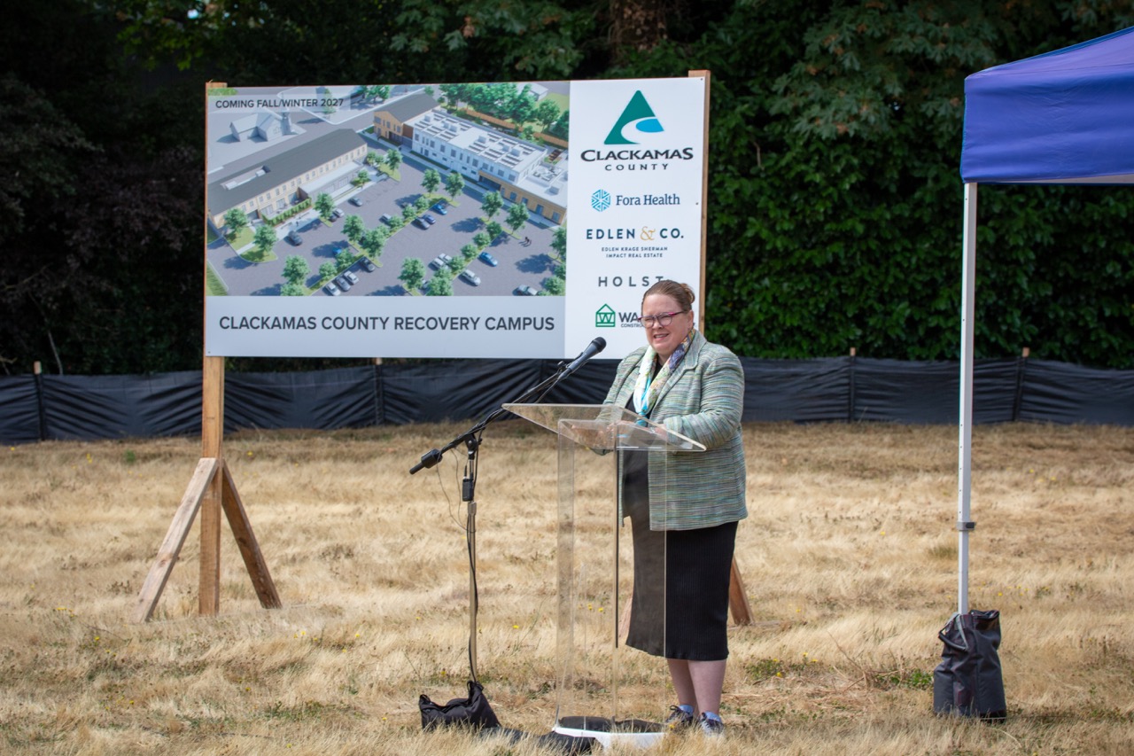 Metro Councilor Christine Lewis speaks behind a lectern at the Clackamas County Recovery Campus groundbreaking on July 29, 2026. In the background is a large sign with a design of the future Clackamas County Recovery Campus.