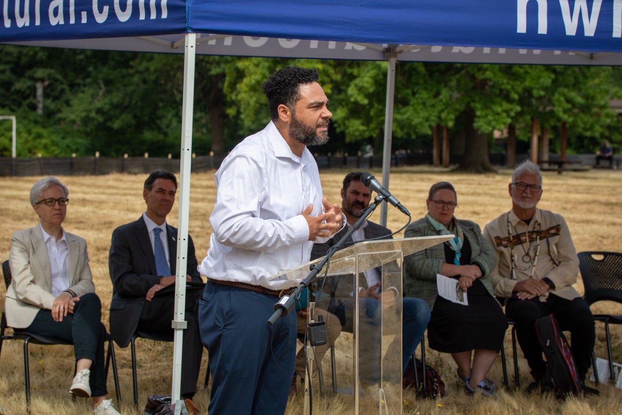 Oregon Recovers' Executive Director Bo Brinson speaks behind a lectern at the Clackamas County Recovery Campus groundbreaking on July 29, 2026. Governor Tina Kotek, Oregon Senate President Rob Wagner, Clackamas County Commissioner Ben West, Metro Councilor Christine Lewis, and Confederated Tribes of Grand Ronde Councilman Jon George are seated in the background.