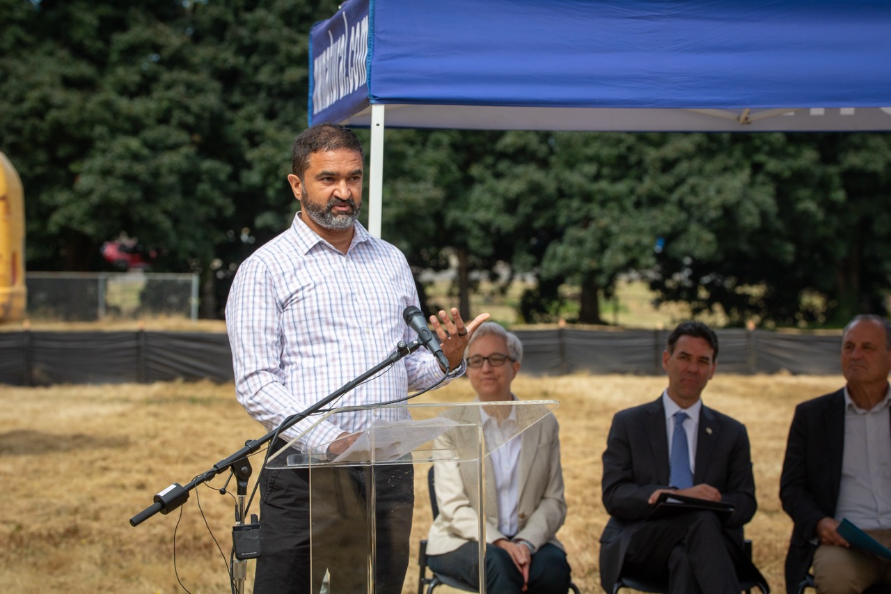 Fora Health CEO Devarshi Bajpai speaks behind a lectern at the Clackamas County Recovery Campus groundbreaking on July 29, 2026. Governor Tina Kotek, Oregon Senate President Rob Wagner, and Clackamas County Chair Craig Roberts are seated in the background.