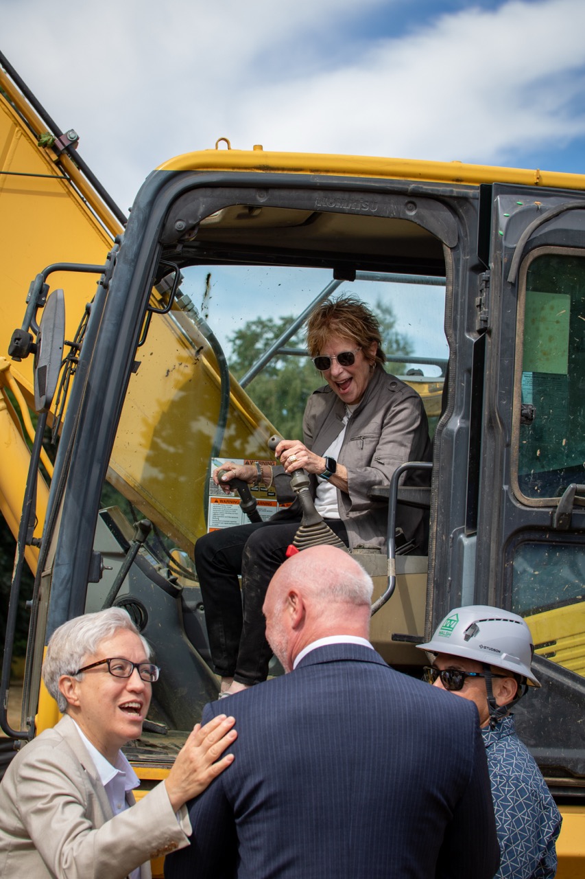 Gov. Tina Kotek laughs with Clackamas County District Attorney John Wentworth at the Clackamas County Recovery Campus groundbreaking on July 29, 2026. A woman is smiling while at the controls of a piece of construction equipment in the background.