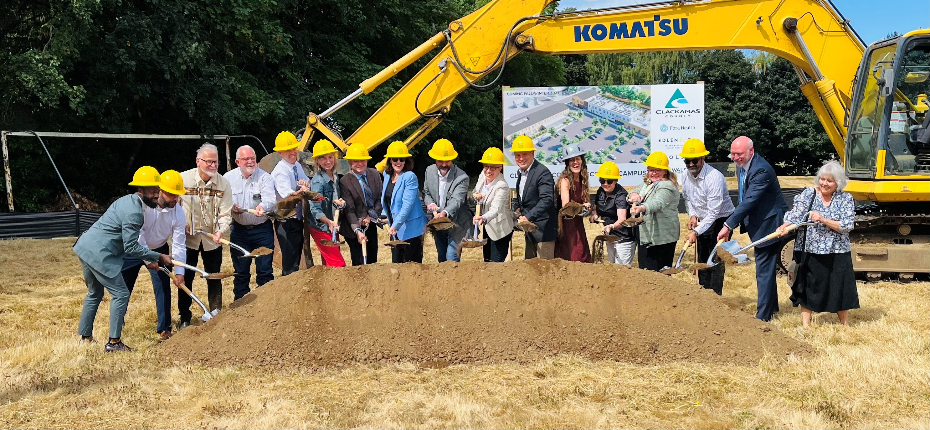 Elected officials shovel a dirt pile with a piece of construction equipment behind them, at the Clackamas County Recovery Campus groundbreaking on July 29, 2026. They are out in a field and all looking up toward a photographer.