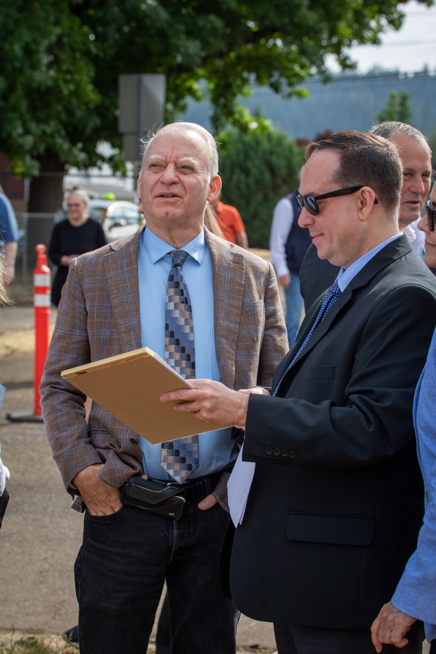 Clackamas County Commissioner Paul Savas and County Administrator Gary Schmidt mingle at the Clackamas County Recovery Campus groundbreaking on July 29, 2026. Savas is looking off in the distance and Schmidt is holding a notepad.