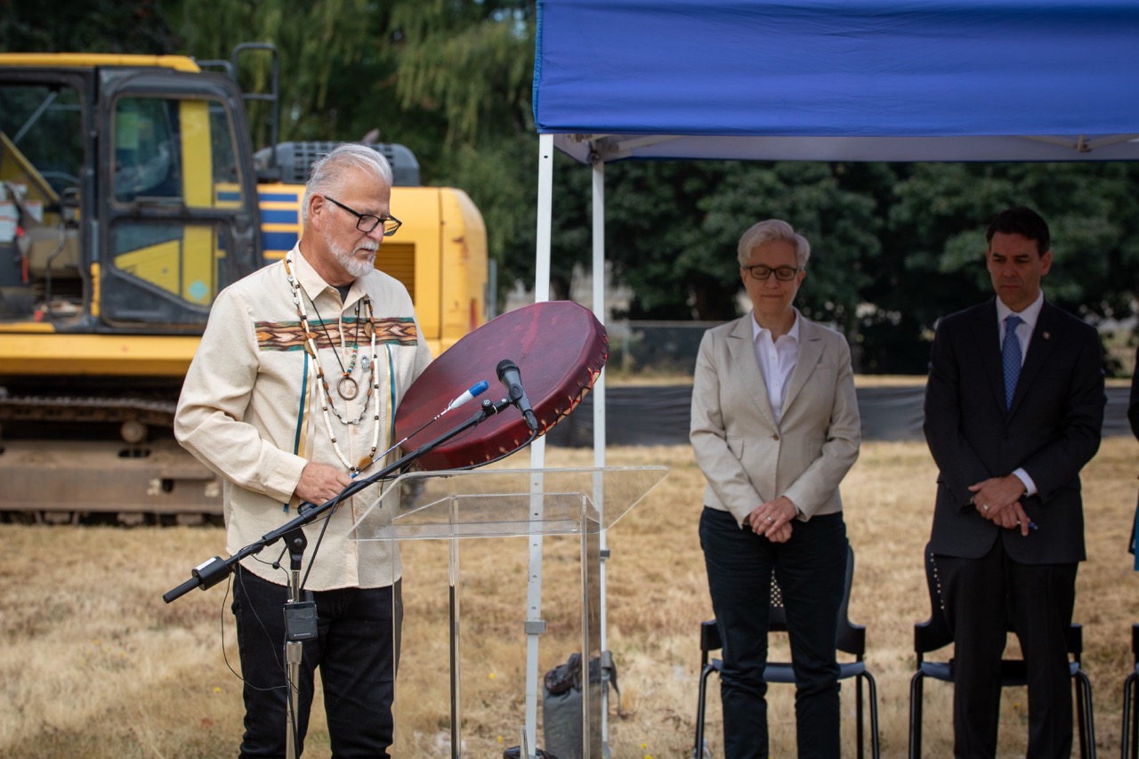 Confederated Tribes of Grand Ronde Councilman Jon George beats a drum as part of a blessing ceremony at the Clackamas County Recovery Campus groundbreaking on July 29, 2026. Gov. Tina Kotek and Oregon Senate President Rob Wagner bow their heads in the background.