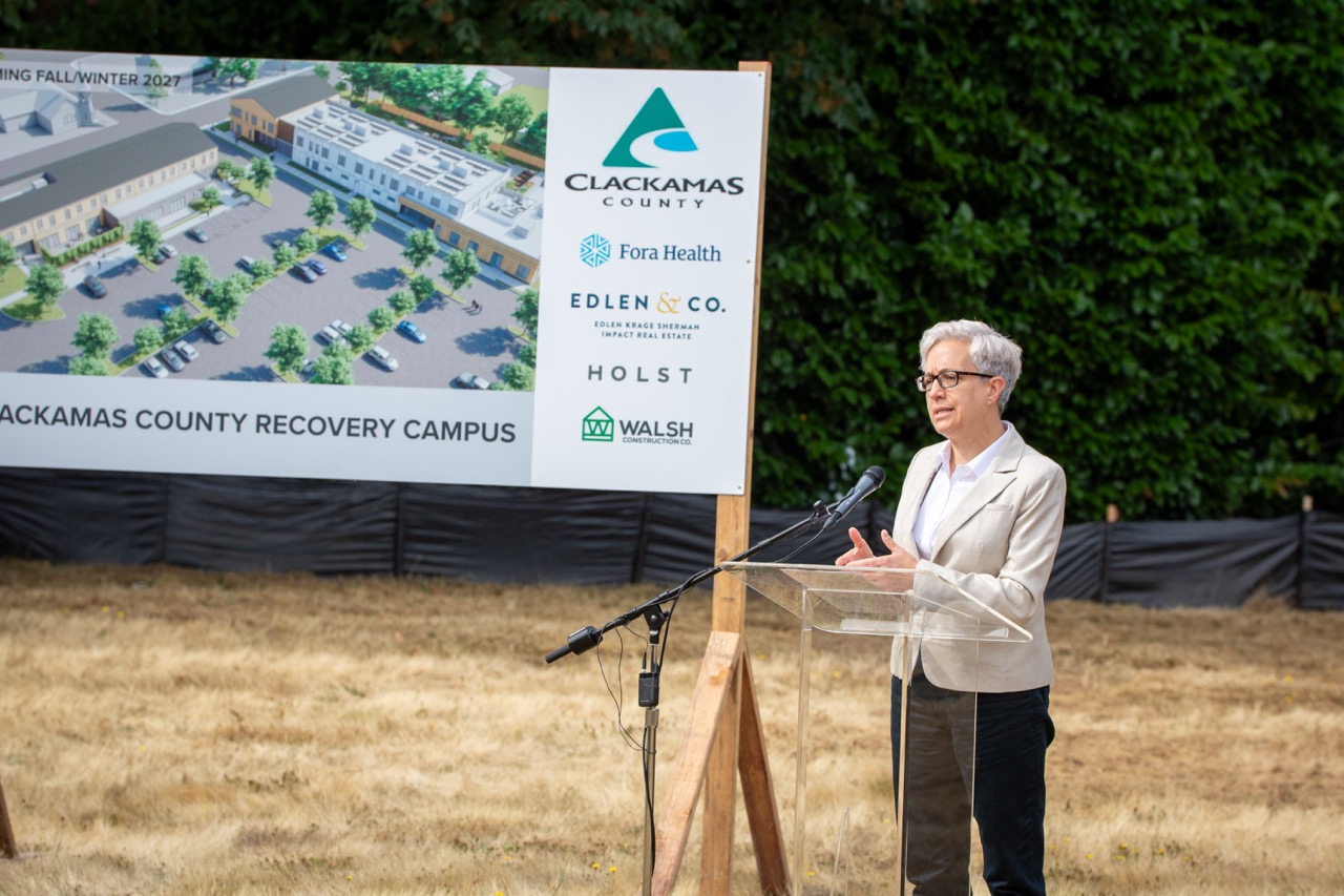 Governor Tina Kotek speaks behind a lectern at the Clackamas County Recovery Campus groundbreaking on July 29, 2026. In the background is a large sign with a design of the future Clackamas County Recovery Campus.