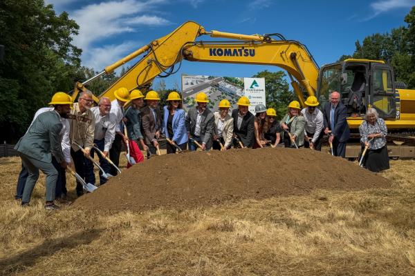 Elected officials shovel a dirt pile with a piece of construction equipment behind them, at the Clackamas County Recovery Campus groundbreaking on July 29, 2026. They are out in a field and all looking up toward a photographer. 