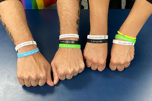 Two people hold out their wrists showing bracelets with encouraging messages that read, "You are not alone," "Are you okay?", and "No estas solo."