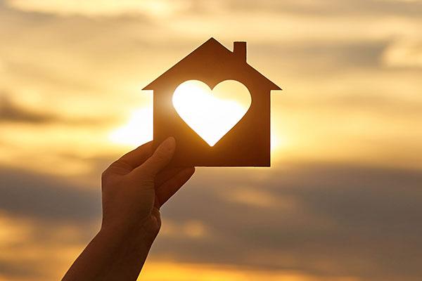 Woman hand holds wooden house in the form of heart against the sun