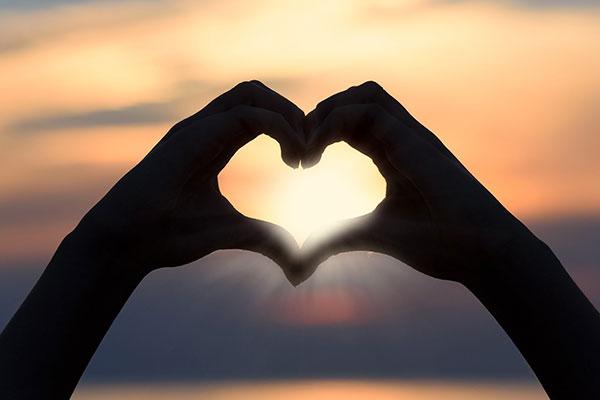 Hands making a heart shape in front of a sun rise/set