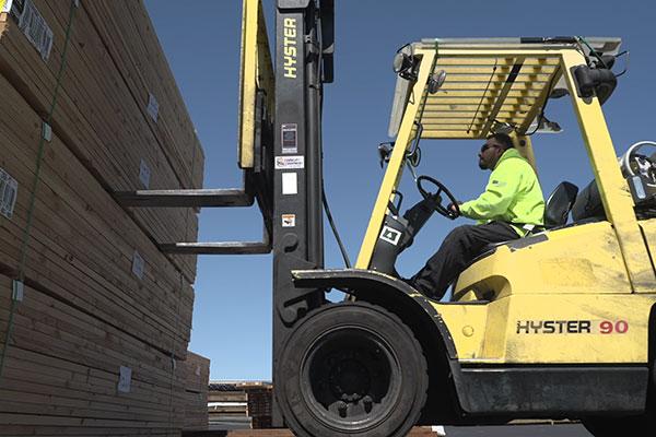 A worker operates a yellow forklift to move a tall stack of wooden planks in an outdoor industrial setting.