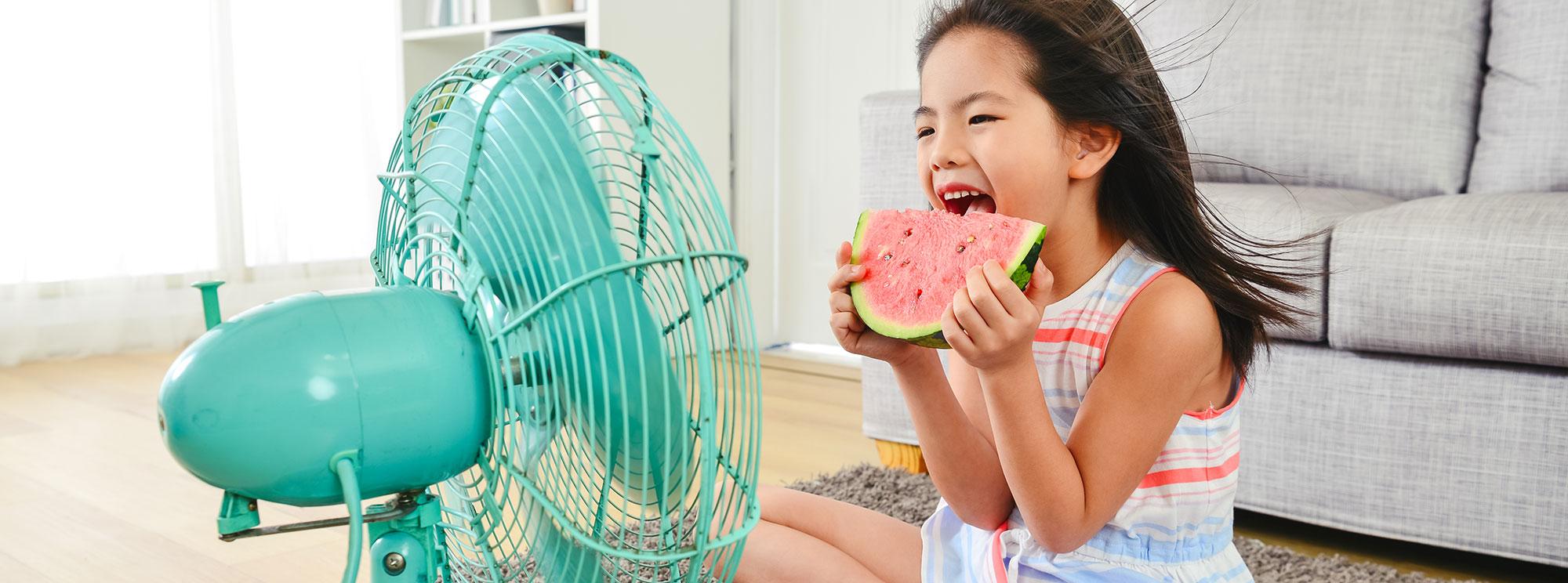 Little girl sitting in front of fan while eating watermelon